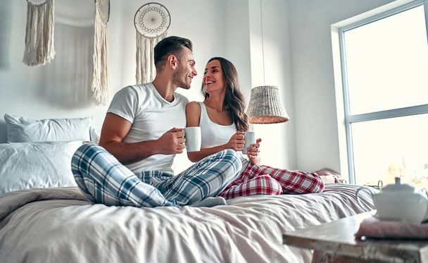 Szczęśliwa młoda para rozmawia i śmieje się siedząc na łóżku i pijąc kawę rano w domu. - Zdjęcie, obraz