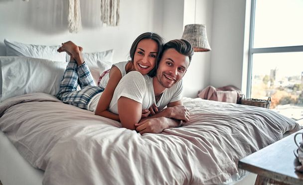 Glückliches junges Paar umarmt und lächelt, während es zu Hause auf dem Bett in einem Schlafzimmer sitzt. - Foto, Bild