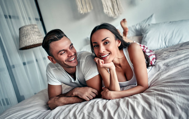 Счастливая молодая пара обнимается и улыбается, сидя на кровати в спальне дома. - Фото, изображение