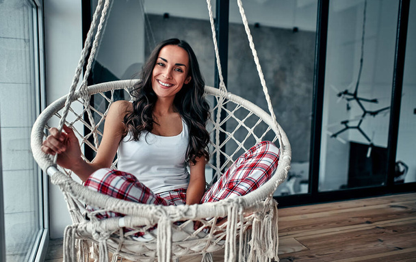 Young woman chilling at home in comfortable hanging chair near the  window. Girl relaxing in swing in loft living room wearing home pajamas. - Foto, imagen