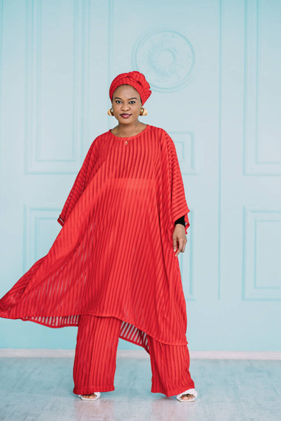 Afrikanisches weibliches Modemodel, das glücklich im Studio posiert. In voller Länge Aufnahme der schönen Frau in stilvollen roten Kleidern stehen über hellblauem Hintergrund. - Foto, Bild