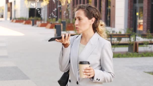Střední pomalý záběr sebevědomé mladé podnikatelky v elegantním kalhotové oblek nahrávání hlasové zprávy nebo mluvení na reproduktoru přes smartphone stojící v centru města s odnést kávu - Záběry, video
