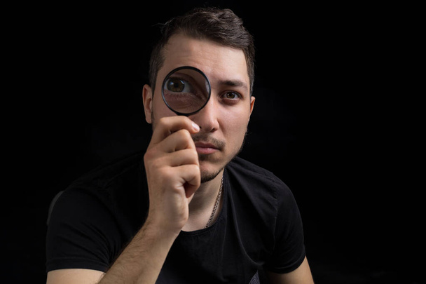 Young handsome man and a magnifying glass on a black background in low key. Search symbol. Investigation concept - Photo, image