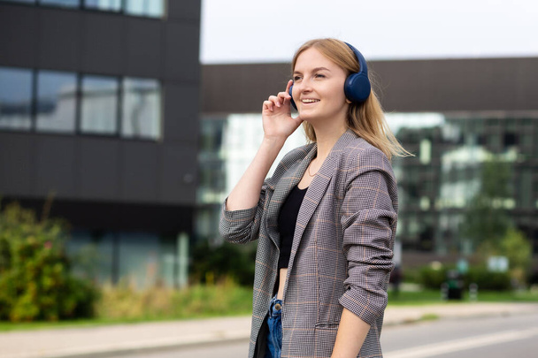 Retrato de una joven mujer feliz escuchando música con auriculares y sonriendo mientras camina por la calle de la ciudad. Amante de la música disfrutando. Retrato de mujer de negocios caminando y sonriendo al aire libre - Foto, imagen