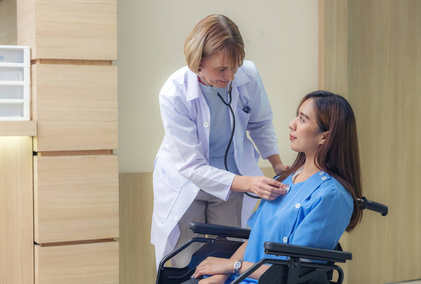 Na chodbě nemocnice, usměvavý lékař vyšetřuje pacienta, asijskou ženu paralyzovanou a sedící na invalidním vozíku. - Fotografie, Obrázek