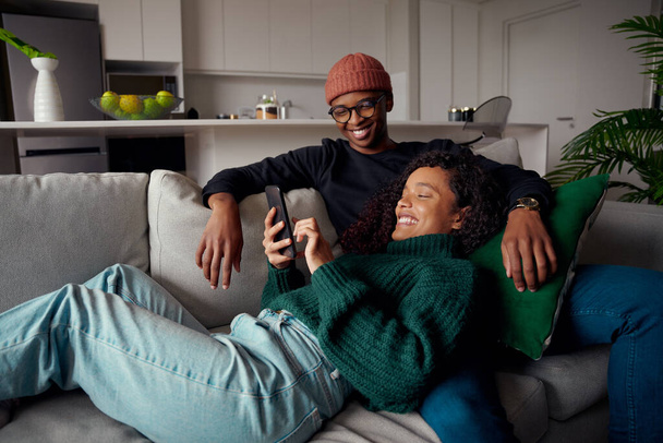 Multi-ethnic couple laughing, looking at text on phone while on the couch in modern apartment - Φωτογραφία, εικόνα