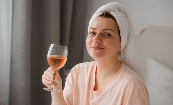 Piękna szczęśliwa młoda kobieta po prysznicu ze szklanką wina w sypialni. Siedzenie w domu w izolatce podczas kwarantanny. Koncepcja świętowania urodzin tylko w łóżku - Zdjęcie, obraz