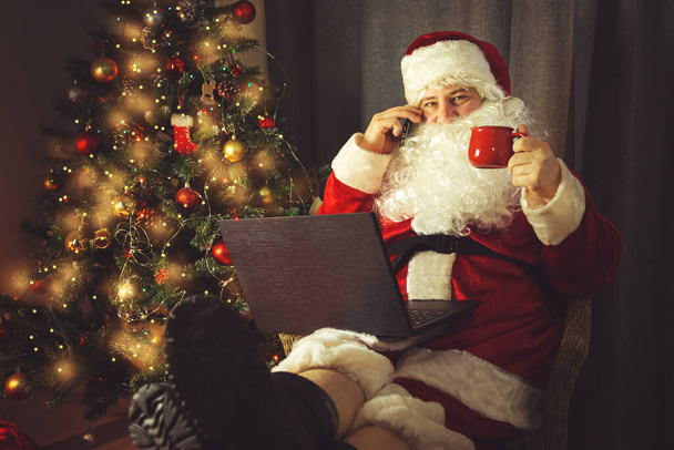 Otantik Noel Baba dizüstü bilgisayar üzerinde çalışıyor ve telefonda konuşuyor. İç mekan. Noel ve Yeni Yıl beklentisiyle.  - Fotoğraf, Görsel