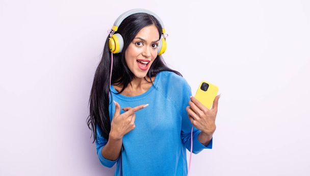pretty hispanic woman looking excited and surprised pointing to the side. smartphone and headphones concept - Foto, Bild