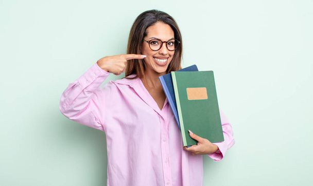 pretty hispanic woman smiling confidently pointing to own broad smile. notebooks concept - Foto, Bild