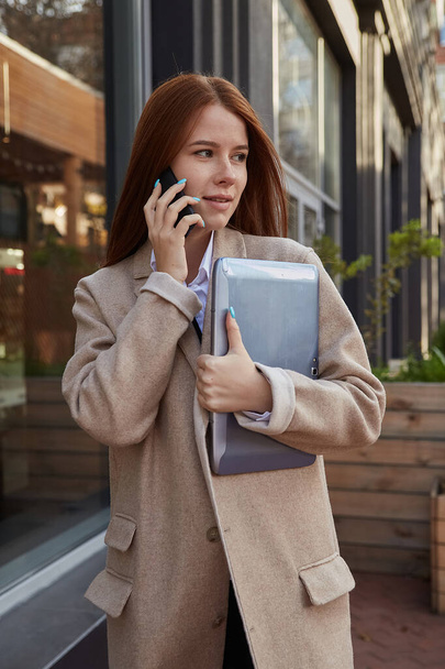 Beyaz kadın şık bej ceketli. Akıllı telefondan arıyor, elinde dizüstü bilgisayar var. - Fotoğraf, Görsel
