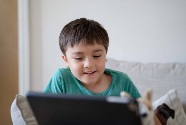 Happy Kid sitzt auf dem Sofa und hat Spaß mit seinen Klassenkameraden auf dem Tablet, Junge lernt online auf dem digitalen Tablet, neuer normaler Lebensstil mit Online-Lernen, Fernunterricht - Foto, Bild
