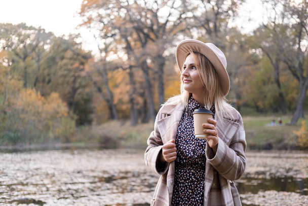 attractive young woman in beige coat and hat drinks coffee by the lake in autumn park in fall. - Zdjęcie, obraz