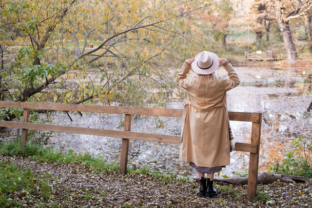 widok z tyłu piękna kobieta w beżowym płaszczu i kapelusz nad jeziorem jesienią parku. - Zdjęcie, obraz