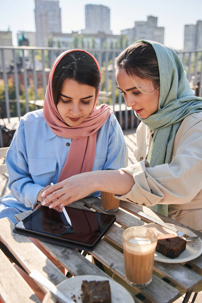 Two arabic sisters in hijab using tablet and drinking coffee together at the street - Valokuva, kuva