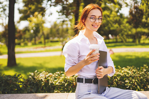 Glad girl student in glasses and white shirt holds laptop and coffee to go while sitting in the park - Foto, imagen