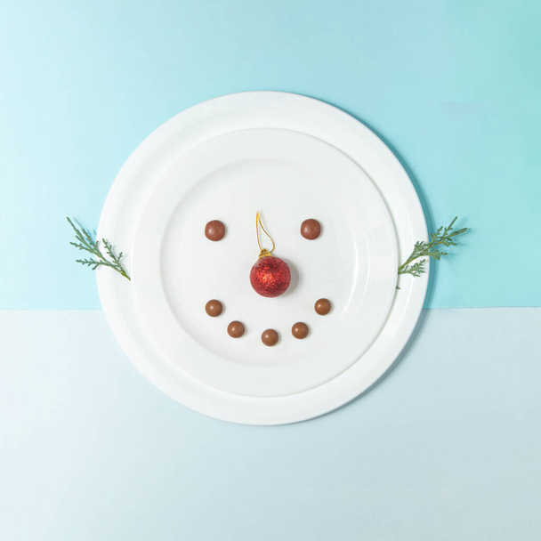 Minimalistic snowman idea. Christmas decoration and chocolate candies in white plates on blue background. Winter concept. New Year flat lay composition. - Photo, Image