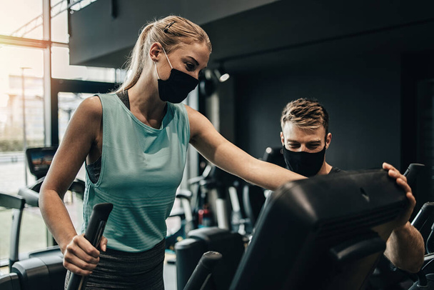 Young fit and attractive woman at body workout in modern gym together with her personal fitness instructor or coach. They keeping distance and wearing protective face masks. Coronavirus world pandemic and sport theme. - Photo, Image