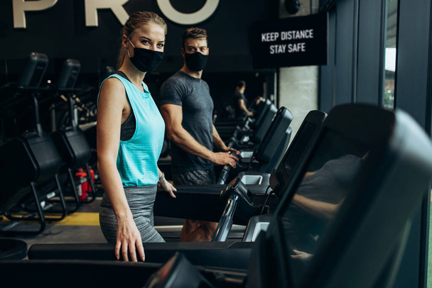 Young fit woman and man running on treadmill in modern fitness gym. They keeping distance and wearing protective face masks. Coronavirus world pandemic and sport theme. - Photo, Image