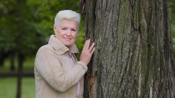 Mutlu, olgun, orta yaşlı, bekar bir kadın kameraya bakıyor. Büyük bir ağacın yanında gülümsüyor. Yaşlı bir kadın. - Video, Çekim