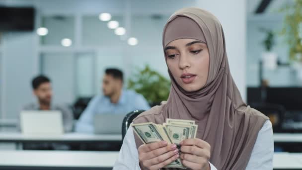 Chica musulmana oriental concentrada grave en hijab gerente oficinista o contable sosteniendo paquete de dólares en las manos contando cuidadosamente dinero en efectivo en el día de pago mantiene registros de los ingresos de la empresa - Imágenes, Vídeo