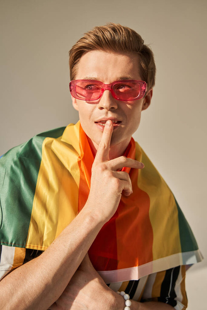 Man in LGBT rainbow flag at shoulders showing silence gesture and gazing sensual away - Fotó, kép