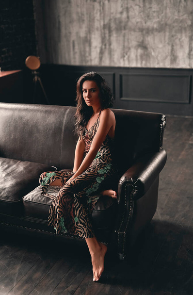 Portrait of a stunning beautiful brunette with curly hair, dressed in a dress that sits on a leather sofa and poses. - Photo, Image