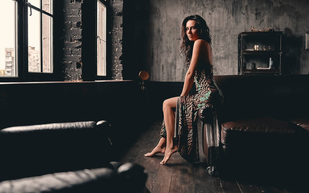Portrait of a stunning beautiful brunette with curly hair, dressed in a dress that sits on a leather sofa and poses, showing long slender legs. - Foto, Imagem