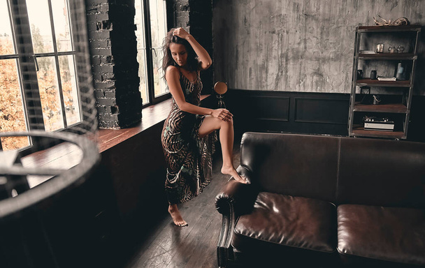 Portrait of a stunning beautiful brunette with curly hair in a dress that poses near a leather sofa and a large window. - Foto, immagini