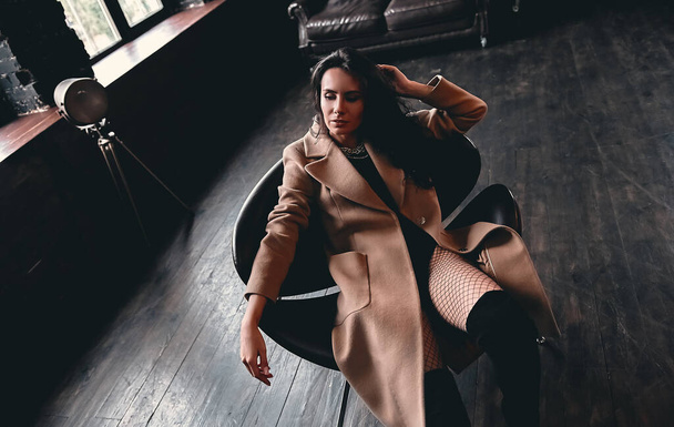 Portrait of a stunning beautiful brunette with curly hair in a coat and mesh tights, who poses on an armchair. - Valokuva, kuva
