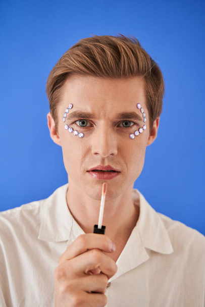 Man holding brush with lips gloss near his face and looking attentively into the lens - Foto, Bild