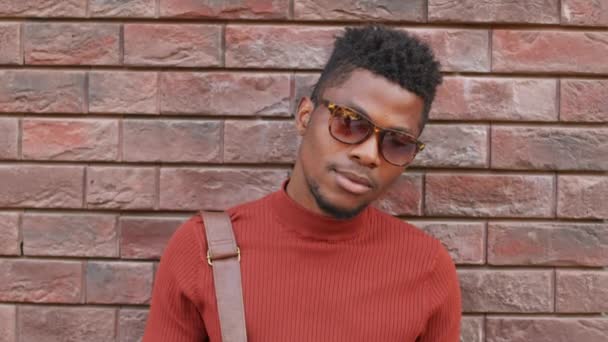 Retrato de primer plano mediano con lentitud de un joven afroamericano guapo con cuello alto rojo y gafas de sol de moda posando para la cámara de pie contra la pared de ladrillo al aire libre - Imágenes, Vídeo
