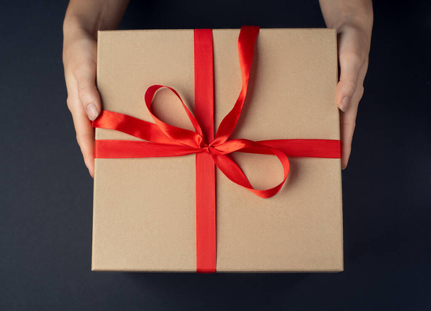 A gift box with a ribbon in woman hands on a dark background - Foto, imagen