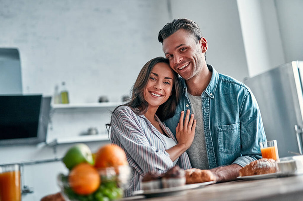Ρομαντικό νεαρό ζευγάρι στην κουζίνα ετοιμάζεται να πάρει πρωινό. Ελκυστική νεαρή γυναίκα και όμορφος άντρας απολαμβάνουν να περνούν χρόνο μαζί ενώ στέκονται στο φως μοντέρνα κουζίνα. - Φωτογραφία, εικόνα