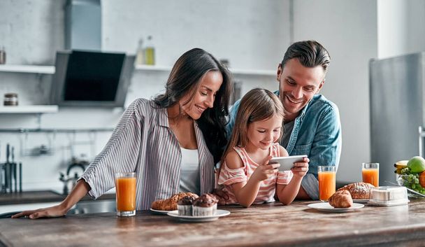 Concepto familiar feliz en la cocina. Linda niña y sus hermosos padres están usando un teléfono inteligente y sonriendo mientras se preparan para un delicioso desayuno. - Foto, imagen