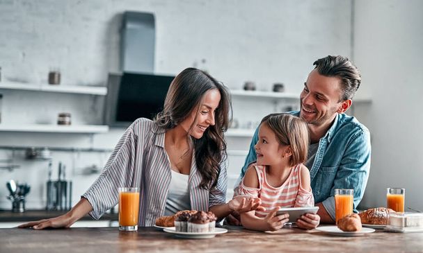 Gelukkig familieconcept in de keuken. Schattig klein meisje en haar mooie ouders zijn met behulp van een smartphone en glimlachen tijdens de voorbereiding voor een heerlijk ontbijt. - Foto, afbeelding