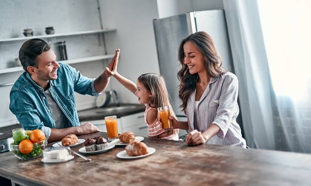 Giving high five to daddy while eating in the kitchen. Happy family are enjoying spending time together while standing on light modern kitchen. - Fotó, kép