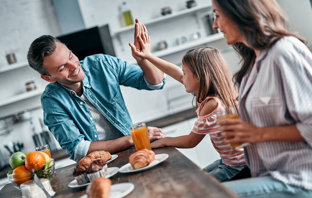 Giving high five to daddy while eating in the kitchen. Happy family are enjoying spending time together while standing on light modern kitchen. - Photo, Image