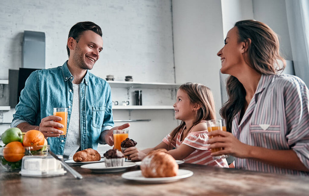 Mama, Papa und ihre kleine schöne Tochter frühstücken in der Küche und unterhalten sich. Glückliches Familienkonzept. - Foto, Bild
