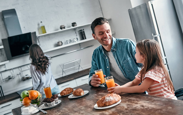 Dad and his little beautiful daughter have breakfast in the kitchen and talk. Mom prepares to eat in the background. Happy family concept. - Photo, Image