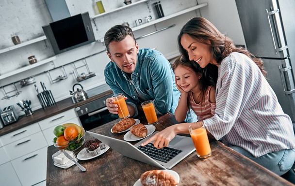 Mama, tata i ich śliczna córeczka jedzą śniadanie w kuchni używając laptopa i rozmawiają. Szczęśliwa koncepcja rodziny. - Zdjęcie, obraz