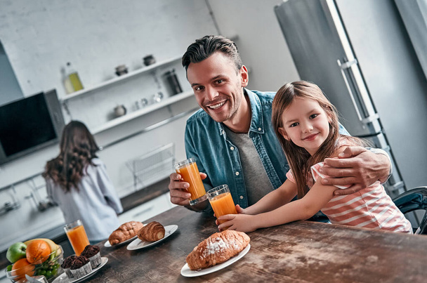 父と彼の小さな美しい娘は台所で朝食を取り、話をします。ママはバックグラウンドで食べる準備ができています。幸せな家庭のコンセプト. - 写真・画像