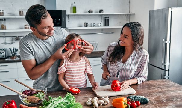 Anya, apa és lánya főznek a konyhában. Boldog családi koncepció. Jóképű férfi, vonzó fiatal nő és az aranyos kislányuk együtt csinálnak salátát. Egészséges életmód. - Fotó, kép