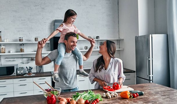 Mami, táta a dcera vaří v kuchyni. Šťastný rodinný koncept. Pohledný muž, atraktivní mladá žena a jejich roztomilá dcerka spolu dělají salát. Zdravý životní styl. - Fotografie, Obrázek