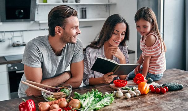 Mom, dad and daughter are cooking on kitchen with recipe book. Happy family concept. Handsome man, attractive young woman and their cute little daughter are making salad together. Healthy lifestyle. - Fotografie, Obrázek