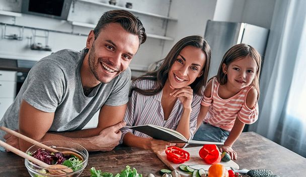 Anya, apa és lánya a konyhában főznek a receptkönyvvel. Boldog családi koncepció. Jóképű férfi, vonzó fiatal nő és az aranyos kislányuk együtt csinálnak salátát. Egészséges életmód. - Fotó, kép
