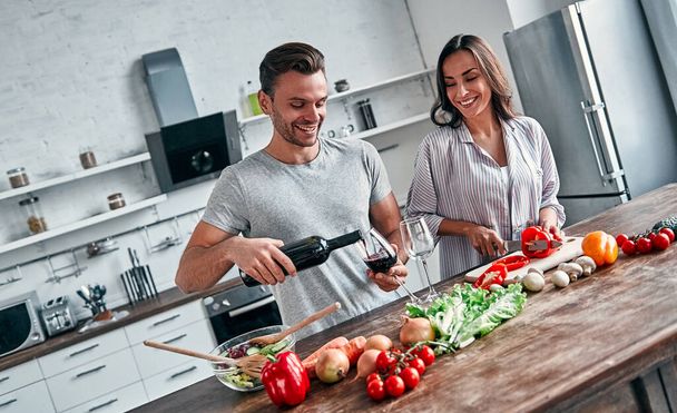 Romantic couple is cooking on kitchen. Handsome man pours wine into glasses and attractive young woman are having fun together while making salad. Healthy lifestyle concept. - 写真・画像