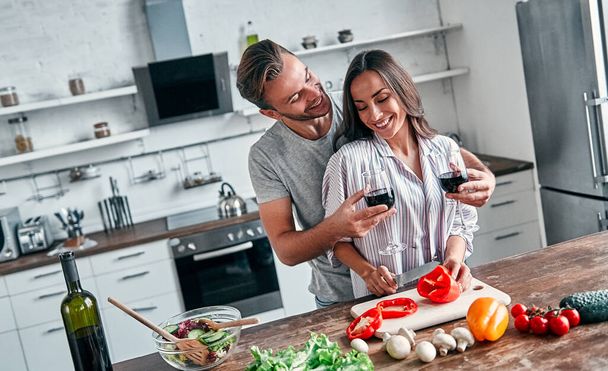 Romantik çift mutfakta yemek pişiriyor. Şarap kadehleriyle yakışıklı bir adam ve çekici genç bir kadın salata yaparken birlikte eğleniyorlar. Sağlıklı yaşam tarzı kavramı. - Fotoğraf, Görsel
