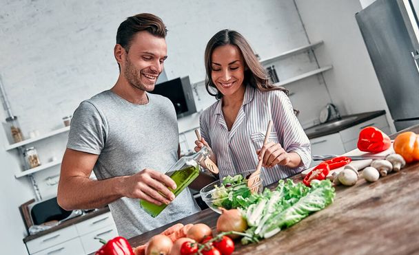 Romantisch koppel kookt in de keuken. Knappe man met een fles olie en aantrekkelijke jonge vrouw hebben plezier samen tijdens het maken van salade. Gezond levensstijl concept. - Foto, afbeelding