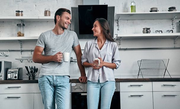 Romantisches junges Paar in der Küche trinkt Kaffee. Attraktive junge Frau und gutaussehender Mann genießen die gemeinsame Zeit, während sie auf der hellen modernen Küche stehen. - Foto, Bild